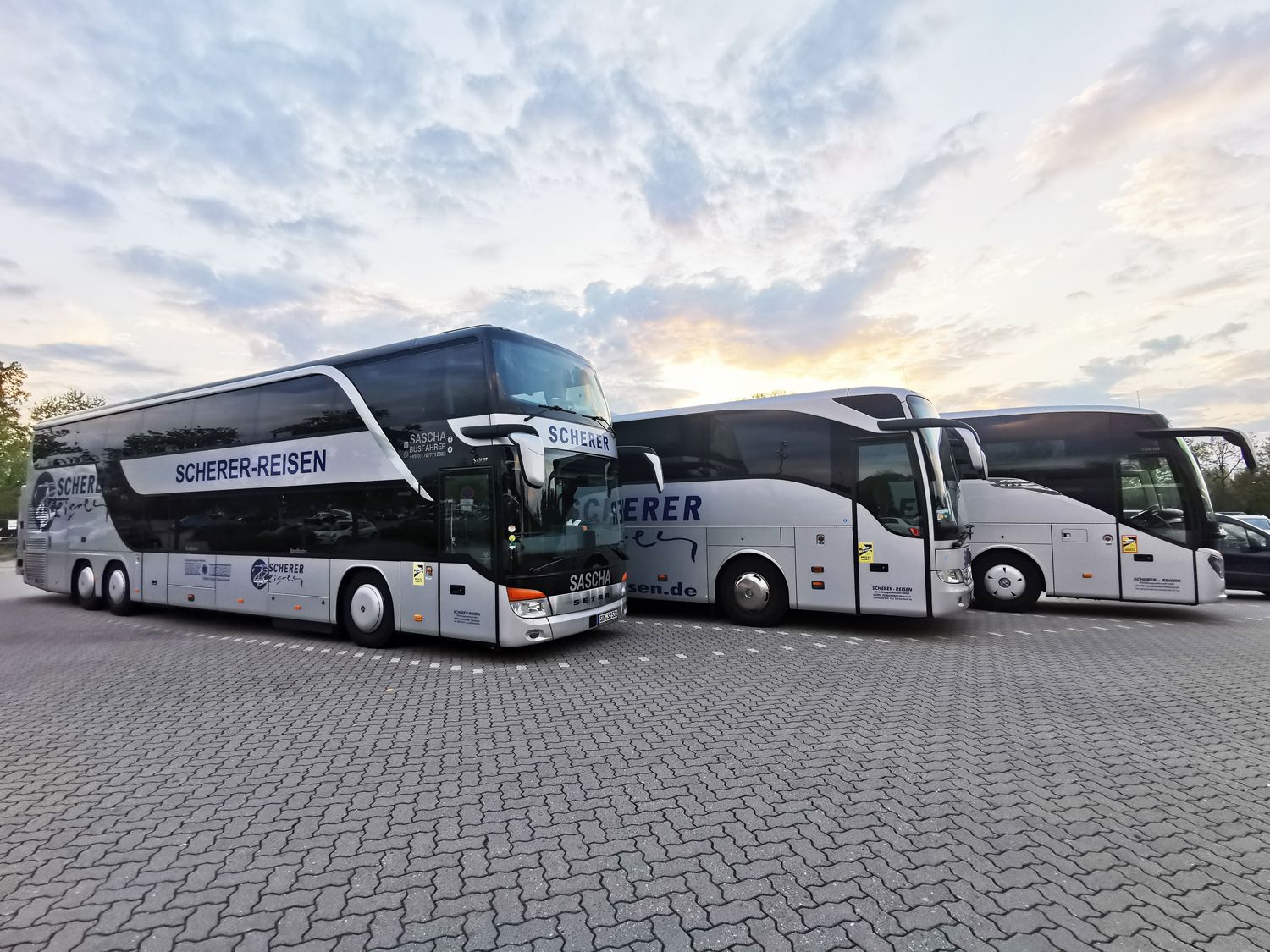 Drei Scherer-Reisen Busse, darunter Saschas silberner Doppeldeckerbus, stehen nebeneinander auf einem Parkplatz.