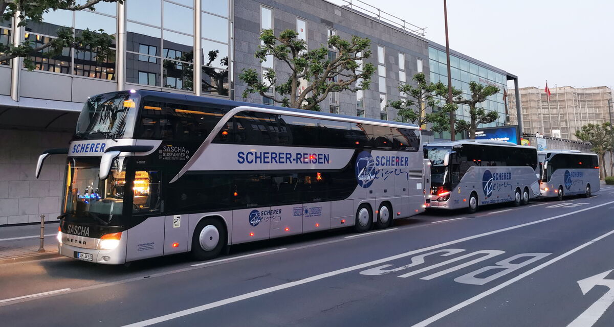 Mehrere Scherer-Reisen Busse stehen hinter Saschas Doppeldeckerbus vor einem Gebäude in einer Stadt.