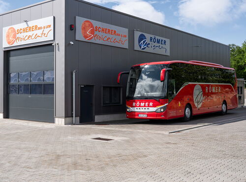 Roter Römer-Reisen Bus vor dem Standort Gebäude Römer-Reisen. Am Gebäude sind Firmenschilder angebracht mit der Aufschrift Scherer Bus & LKW Servicecenter und "Römer Reisen.