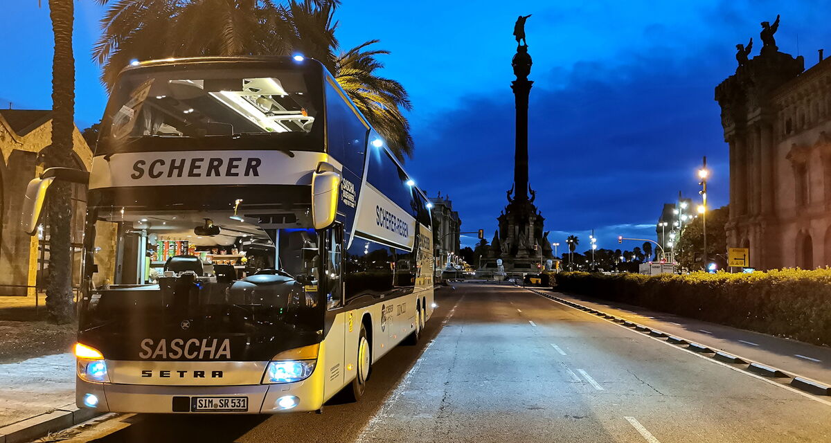 Saschas silberner Doppeldeckerbus vor dem Kolumbus-Denkmal in Barcelona auf einer Straße in der Dämmerung mit Palmen.