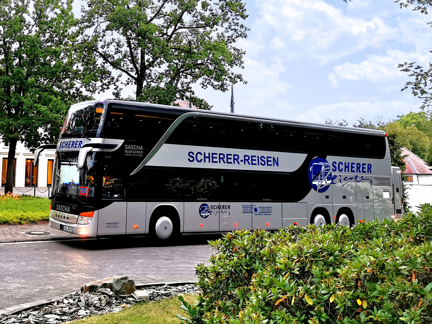 Saschas silberner Doppeldeckerbus auf einer Straße zwischen Bäumen und Sträuchern.