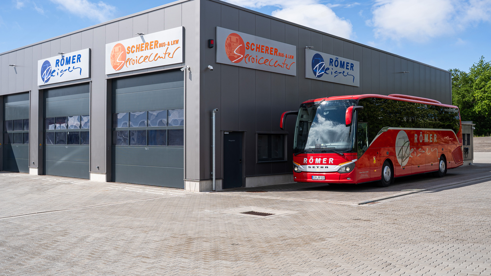 Roter Römer-Reisen Bus vor dem Standort Gebäude Römer-Reisen. Am Gebäude sind Firmenschilder angebracht mit der Aufschrift Scherer Bus & LKW Servicecenter und "Römer Reisen.